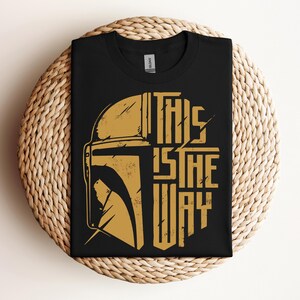 May include: Black t-shirt featuring a gold-colored illustration of a helmet and the phrase "THIS IS THE WAY." The design is a stylized, distressed graphic. The shirt is folded and placed on a woven, circular surface.