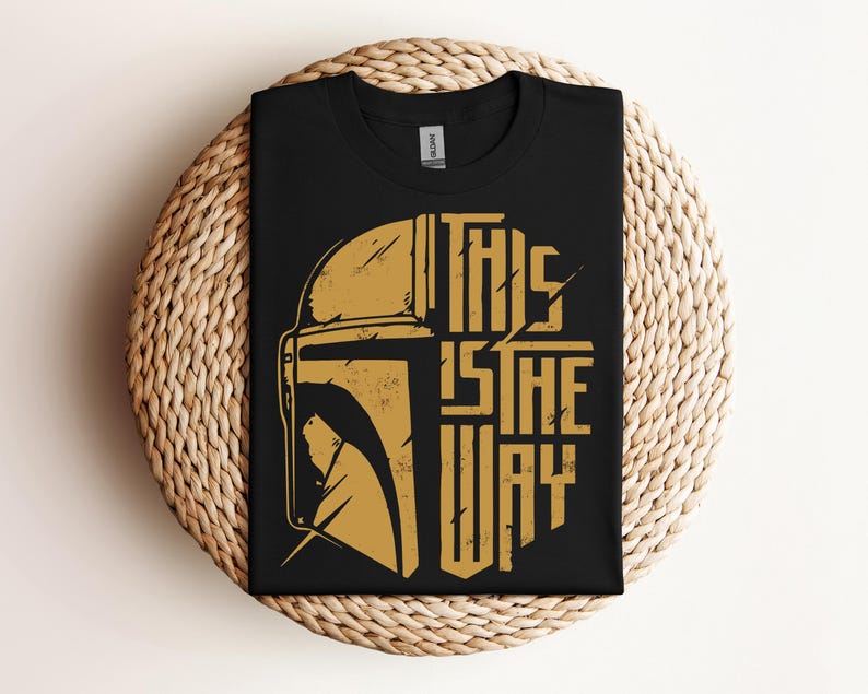 May include: Black t-shirt featuring a gold-colored illustration of a helmet and the phrase "THIS IS THE WAY." The design is a stylized, distressed graphic. The shirt is folded and placed on a woven, circular surface.