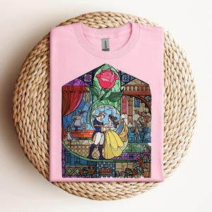 May include: Pink t-shirt featuring a stained-glass design with a rose and characters dancing. The design includes a yellow dress, blue suit, and other figures. The shirt is folded on a woven, circular surface.