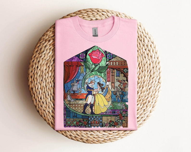 May include: Pink t-shirt featuring a stained-glass design with a rose and characters dancing. The design includes a yellow dress, blue suit, and other figures. The shirt is folded on a woven, circular surface.