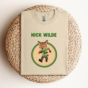 May include: Beige t-shirt featuring the name "NICK WILDE" in green block letters above a cartoon fox character inside a green and yellow circle. The fox is wearing a green shirt and tie.