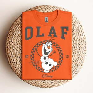 Może przedstawiać: Pomarańczowy T-shirt z grafiką Olafa z Krainy Lodu. Grafika przedstawia Olafa w k&oacute;łku z kratkowanym obramowaniem i tekstem "Olaf" nad grafiką. Tekst "Disney" znajduje się pod grafiką.