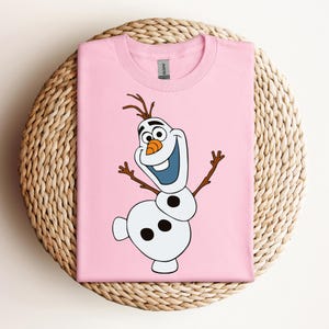 Pode incluir: Camiseta rosa com uma imagem de desenho animado de Olaf do filme Frozen. Olaf &eacute; um boneco de neve com um nariz de cenoura, olhos pretos e um grande sorriso. Ele usa um chap&eacute;u de galho de &aacute;rvore marrom e tem bra&ccedil;os de galho de &aacute;rvore marrom.