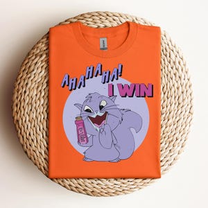 Puede incluir: Camiseta naranja con un gato morado de dibujos animados sosteniendo un objeto rosa y el texto "AHA HA HA! I WIN".