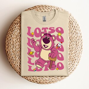 Puede incluir: Una camiseta beige con un oso de dibujos animados rosa que lleva una camisa morada y sostiene un mazo de madera. El oso est&aacute; rodeado de la palabra "LOTSO" en una fuente retro rosa con estrellas y otros gr&aacute;ficos de tem&aacute;tica espacial.