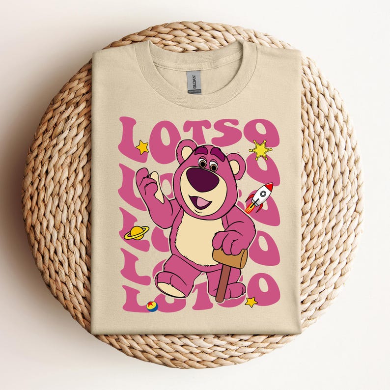 Puede incluir: Una camiseta beige con un oso de dibujos animados rosa que lleva una camisa morada y sostiene un mazo de madera. El oso est&aacute; rodeado de la palabra "LOTSO" en una fuente retro rosa con estrellas y otros gr&aacute;ficos de tem&aacute;tica espacial.