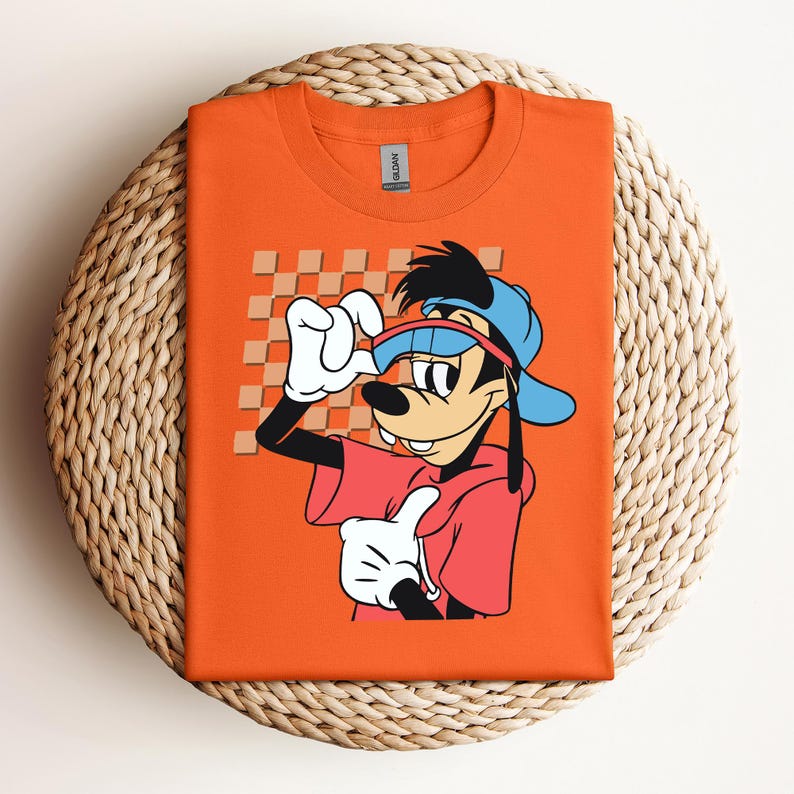 May include: Orange t-shirt featuring a cartoon illustration of Goofy wearing sunglasses and a blue baseball cap. Goofy is wearing a red shirt with a white checkerboard pattern in the background.