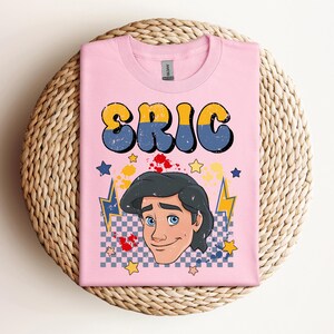 May include: Pink t-shirt featuring the name "ERIC" in yellow and blue bubble letters. Below the name is a cartoon illustration of a man with dark hair and blue eyes. The design includes stars, lightning bolts, and a checkered pattern.