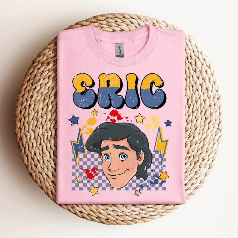 May include: Pink t-shirt featuring the name "ERIC" in yellow and blue bubble letters. Below the name is a cartoon illustration of a man with dark hair and blue eyes. The design includes stars, lightning bolts, and a checkered pattern.