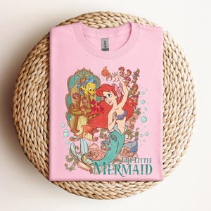 May include: Pink t-shirt featuring a colorful graphic of Ariel from The Little Mermaid. The design includes Ariel, Flounder, and other sea creatures, with the words "The Little Mermaid" in teal.