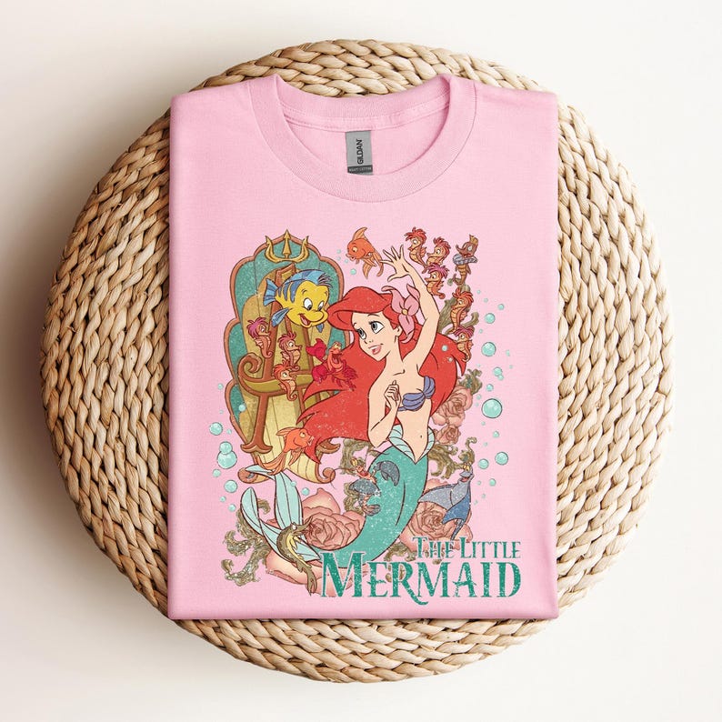 May include: Pink t-shirt featuring a colorful graphic of Ariel from The Little Mermaid. The design includes Ariel, Flounder, and other sea creatures, with the words "The Little Mermaid" in teal.