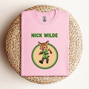 May include: A light pink t-shirt featuring the text "NICK WILDE" in green above a cartoon image of a fox character in a green circle. The fox is wearing a green shirt and tie. The t-shirt is folded on a woven, circular mat.