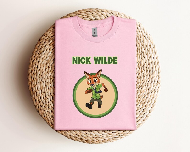 May include: A light pink t-shirt featuring the text "NICK WILDE" in green above a cartoon image of a fox character in a green circle. The fox is wearing a green shirt and tie. The t-shirt is folded on a woven, circular mat.