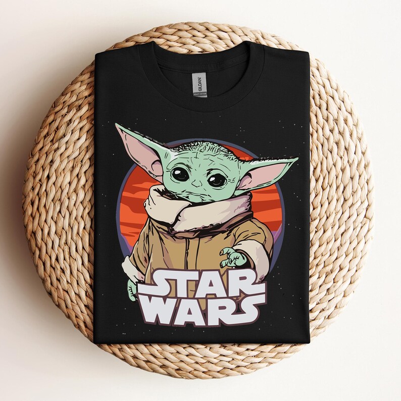 Op de afbeelding: Zwart T-shirt met een afbeelding van een groen alien-personage met grote oren, tegen een rode en oranje achtergrond. De woorden "STAR WARS" staan eronder in het wit gedrukt.