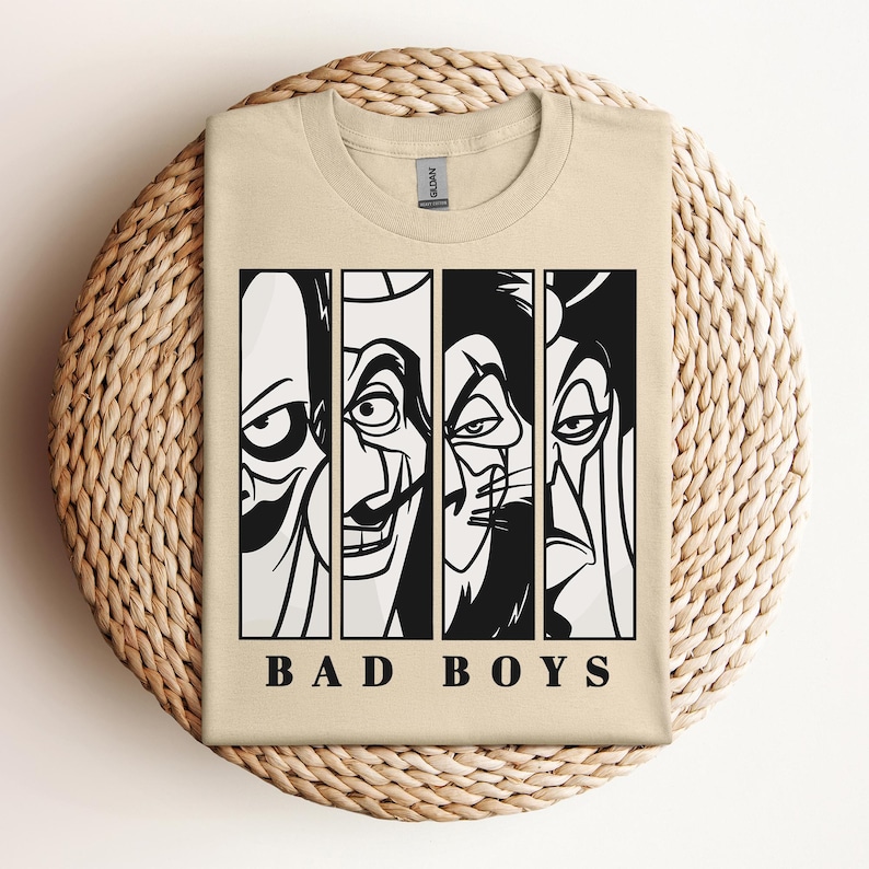 May include: A tan t-shirt featuring a graphic of four villainous faces in black and white, with the words "BAD BOYS" printed below. The shirt is folded on a woven, circular surface.
