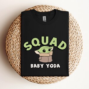 May include: Black t-shirt featuring a green and white graphic of Baby Yoda with the word "SQUAD" in green and white, and the words "BABY YODA" in white. The t-shirt is displayed on a woven, circular surface.