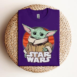 Op de afbeelding: Een paars T-shirt met een afbeelding van een groen alien-personage met grote oren, tegen een oranje en rode achtergrond. De woorden "STAR WARS" zijn in het wit onder het personage gedrukt. Het shirt wordt gepresenteerd op een geweven, cirkelvormig oppervlak.