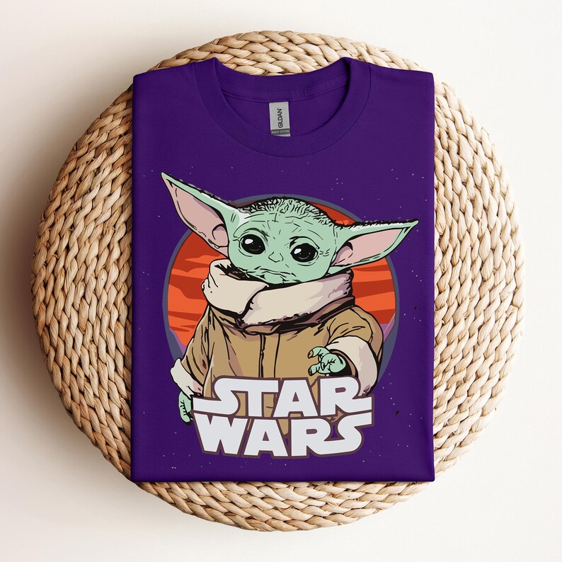 Op de afbeelding: Een paars T-shirt met een afbeelding van een groen alien-personage met grote oren, tegen een oranje en rode achtergrond. De woorden "STAR WARS" zijn in het wit onder het personage gedrukt. Het shirt wordt gepresenteerd op een geweven, cirkelvormig oppervlak.