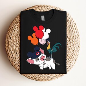 Op de afbeelding: Zwart T-shirt met een cartoonvarken met een marineblauwe hoed en ballonnen. Een haan staat op de rug van het varken. De ballonnen zijn rood, wit en oranje. Het shirt ligt op een geweven oppervlak.