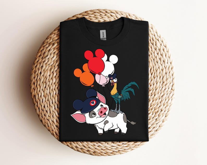 Op de afbeelding: Zwart T-shirt met een cartoonvarken met een marineblauwe hoed en ballonnen. Een haan staat op de rug van het varken. De ballonnen zijn rood, wit en oranje. Het shirt ligt op een geweven oppervlak.