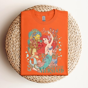 May include: Orange t-shirt featuring a graphic of Ariel from The Little Mermaid. The design includes Ariel with red hair, a blue seashell bra, and a teal mermaid tail, along with Flounder and other sea creatures. The words "Little Mermaid" are printed in teal.