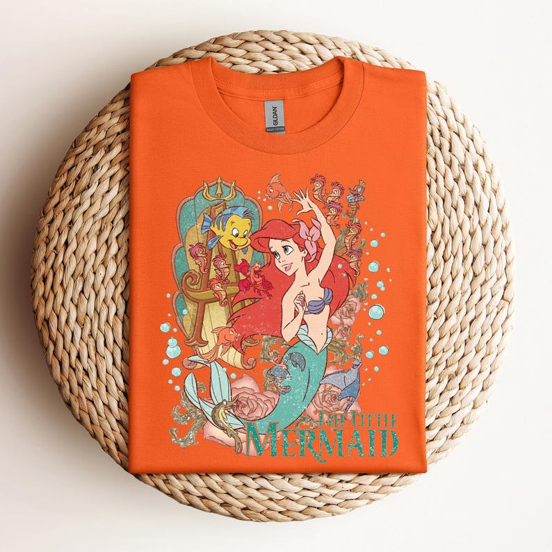 May include: Orange t-shirt featuring a graphic of Ariel from The Little Mermaid. The design includes Ariel with red hair, a blue seashell bra, and a teal mermaid tail, along with Flounder and other sea creatures. The words "Little Mermaid" are printed in teal.