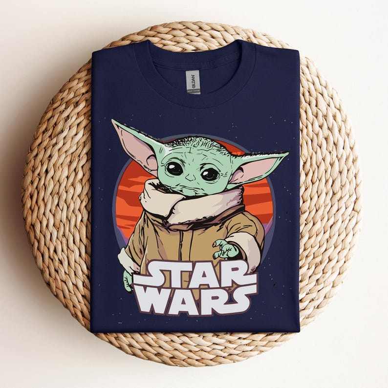 Op de afbeelding: Marineblauw T-shirt met een afbeelding van een groen alien-personage met grote oren, tegen een rode en oranje achtergrond. De woorden "STAR WARS" zijn in het wit onder het personage gedrukt. Het T-shirt wordt gepresenteerd op een geweven oppervlak.
