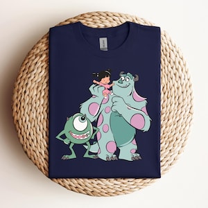 May include: A navy blue t-shirt with a graphic of Mike and Sulley from Monsters Inc. and Boo. Mike is green with one large eye and Sulley is blue with pink spots. Boo is wearing a pink dress and is being held by Sulley.
