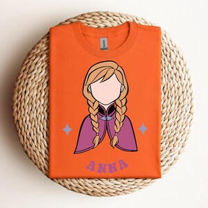 May include: An orange t-shirt featuring a graphic illustration of a character with blonde hair in two braids, wearing a purple cape. The character has no facial features. Below the graphic, the name ANNA is printed in purple letters.