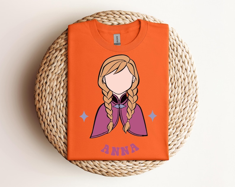 May include: An orange t-shirt featuring a graphic illustration of a character with blonde hair in two braids, wearing a purple cape. The character has no facial features. Below the graphic, the name ANNA is printed in purple letters.