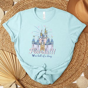 Puede incluir: Camiseta azul claro con una ilustraci&oacute;n colorida de un castillo y el texto "Florida!!!" y "one hell of a drug." El dise&ntilde;o incluye fuegos artificiales y corazones. La camiseta est&aacute; hecha de un material suave.
