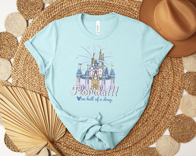 Puede incluir: Camiseta azul claro con una ilustraci&oacute;n colorida de un castillo y el texto "Florida!!!" y "one hell of a drug." El dise&ntilde;o incluye fuegos artificiales y corazones. La camiseta est&aacute; hecha de un material suave.