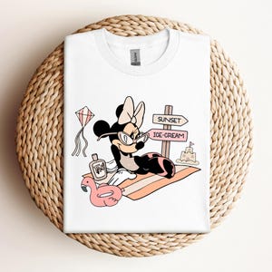 K&ouml;nnte beinhalten: Wei&szlig;es T-Shirt mit einem Cartoon-Bild von Minnie Mouse, die eine Sonnenbrille und eine Schleife tr&auml;gt und sich auf einem Strandtuch mit einem rosafarbenen Flamingo-Schwimmring, Sonnencreme und einem Schild mit der Aufschrift "Sunset" und "Ice-Cream" entspannt.
