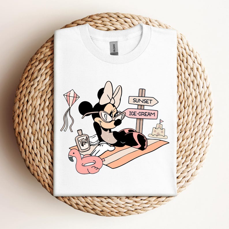 K&ouml;nnte beinhalten: Wei&szlig;es T-Shirt mit einem Cartoon-Bild von Minnie Mouse, die eine Sonnenbrille und eine Schleife tr&auml;gt und sich auf einem Strandtuch mit einem rosafarbenen Flamingo-Schwimmring, Sonnencreme und einem Schild mit der Aufschrift "Sunset" und "Ice-Cream" entspannt.