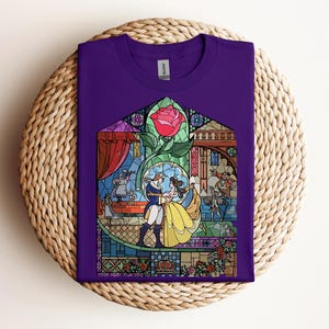 May include: A purple t-shirt featuring a stained-glass design with characters from a fairy tale. The design includes a red rose, a dancing couple, and other figures in a colorful, detailed illustration. The shirt is folded on a woven surface.