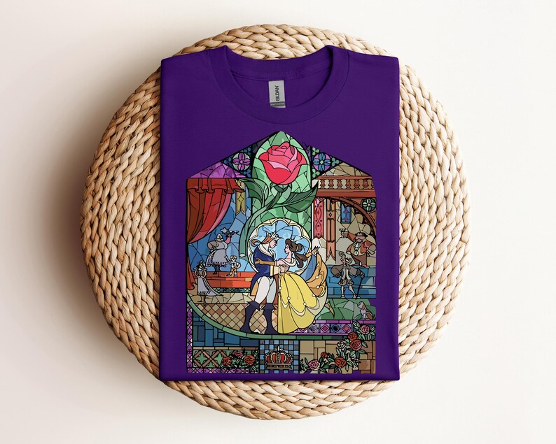 May include: A purple t-shirt featuring a stained-glass design with characters from a fairy tale. The design includes a red rose, a dancing couple, and other figures in a colorful, detailed illustration. The shirt is folded on a woven surface.