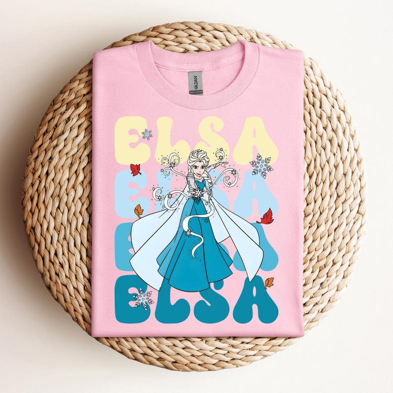 Op de afbeelding: Roze T-shirt met een afbeelding van Elsa uit Frozen. Het woord "ELSA" staat in een retro lettertype, lichtblauw en geel. Het ontwerp bevat sneeuwvlokken en herfstbladeren.