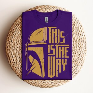 May include: Purple t-shirt with a gold graphic of a helmet and the text "THIS IS THE WAY." The shirt is folded on a round, woven, beige placemat. The shirt has a crew neck and a tag.