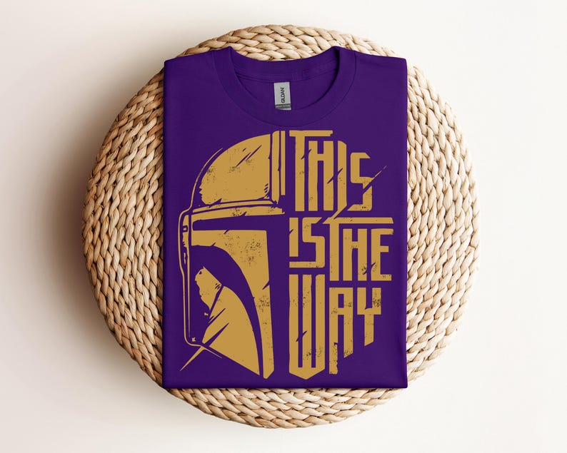 May include: Purple t-shirt with a gold graphic of a helmet and the text "THIS IS THE WAY." The shirt is folded on a round, woven, beige placemat. The shirt has a crew neck and a tag.