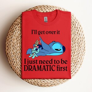 Puede incluir: Camiseta roja con un personaje azul Stitch de Lilo & Stitch. Stitch est&aacute; acostado con una peque&ntilde;a mu&ntilde;eca. El texto en la camiseta dice "I'll get over it I just need to be DRAMATIC first."