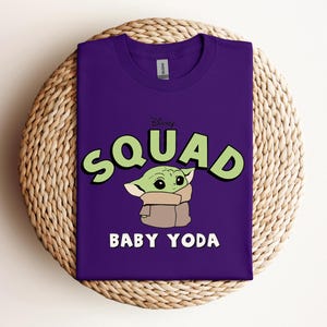 May include: A purple t-shirt featuring the text "SQUAD" in green letters with a black outline, and a cartoon image of Baby Yoda. The text "BABY YODA" is below the image in white letters. The shirt is displayed on a woven, circular surface.