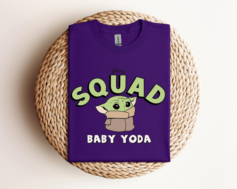 May include: A purple t-shirt featuring the text "SQUAD" in green letters with a black outline, and a cartoon image of Baby Yoda. The text "BABY YODA" is below the image in white letters. The shirt is displayed on a woven, circular surface.