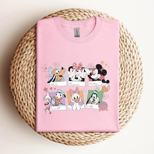 May include: A light pink short-sleeved t-shirt featuring a graphic print of six individual polaroid-style frames. Each frame contains a different Disney character: Pluto, Minnie Mouse, Mickey Mouse, Donald Duck, Daisy Duck, and Goofy.