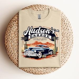 May include: A tan t-shirt featuring a vintage car graphic with the text "Hudson's Garage" in a retro design. The graphic includes a classic blue car, cacti, and mountain scenery, with the text "Serving the Springs Since 1955."