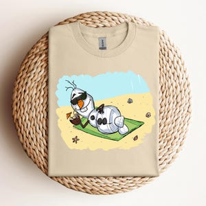 Op de afbeelding: Een beige T-shirt met een cartoon afbeelding van Olaf uit Frozen, die een zonnebril draagt en ontspant op een strand met een kokosnootdrankje. De afbeelding is op een lichtbeige achtergrond.