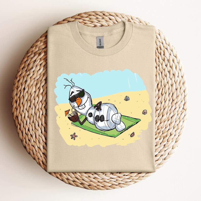 Op de afbeelding: Een beige T-shirt met een cartoon afbeelding van Olaf uit Frozen, die een zonnebril draagt en ontspant op een strand met een kokosnootdrankje. De afbeelding is op een lichtbeige achtergrond.