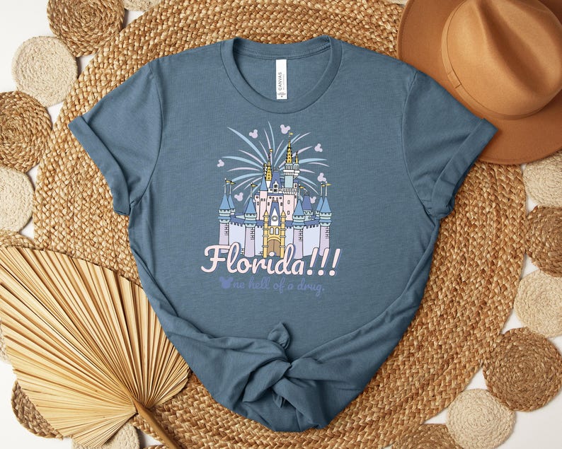 Puede incluir: Una camiseta azul con un castillo, fuegos artificiales y corazones. La palabra "Florida!!!" est&aacute; escrita en una fuente caprichosa, con la frase "no hell of a drug" debajo. La camiseta est&aacute; hecha de un material suave.