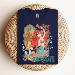 May include: Navy blue t-shirt featuring a colorful graphic of Ariel from The Little Mermaid. The design includes Ariel with red hair, a turquoise tail, and a seashell bra, along with Flounder and other sea creatures. The words "The Little Mermaid" are printed below.