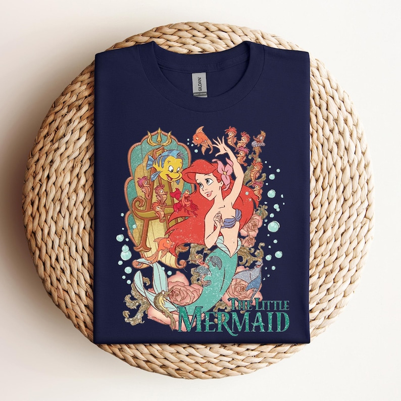 May include: Navy blue t-shirt featuring a colorful graphic of Ariel from The Little Mermaid. The design includes Ariel with red hair, a turquoise tail, and a seashell bra, along with Flounder and other sea creatures. The words "The Little Mermaid" are printed below.