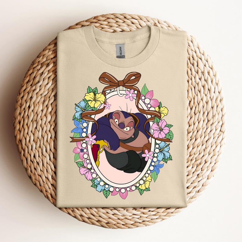 Puede incluir: Camiseta beige con un personaje de dibujos animados en un marco floral ovalado. El personaje es azul oscuro y negro, con una expresi&oacute;n traviesa. El marco incluye una cinta marr&oacute;n y flores rosas, amarillas y azules.